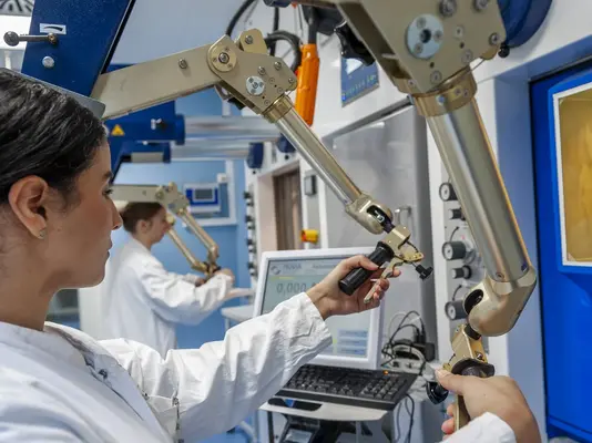 Eine Laborantin bedient einen mechanischen Arm in einem modernen Forschungslabor. Sie trägt einen weißen Laborkittel und konzentriert sich auf ihre Arbeit. Im Hintergrund sind weitere Forscher und Laborausrüstung sichtbar. Die Aufnahme vermittelt ein Gefühl von Wissenschaft und technologischem Fortschritt.