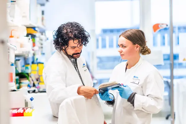 Zwei Wissenschaftler in Laborkitteln betrachten zusammen ein Notizbuch in einem modernen Labor. Der Mann mit lockigen Haaren zeigt auf eine Seite, während die Frau, die Gummihandschuhe trägt, aufmerksam schaut. Im Hintergrund sind Labormaterialien sichtbar.