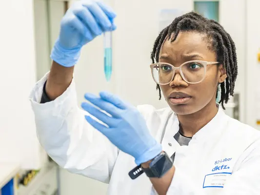 Eine Wissenschaftlerin in einem Laborkittel untersucht aufmerksam eine Pipette mit blauem Inhalt. Sie trägt Schutzbrille und Handschuhe, während sie die Probe in einem modernen Labor betrachtet. Hintergrund mit Laborequipment und neutralen Farben.