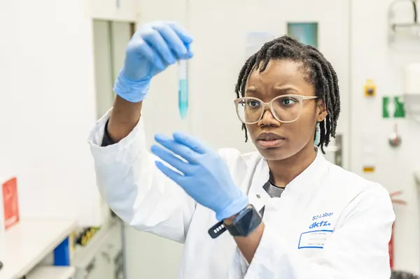 A focused laboratory technician, wearing a white coat and gloves, carefully examines a test tube with a blue liquid. She appears thoughtful and concerned, suggesting she is analyzing the contents for research purposes. The laboratory setting includes scientific equipment in the background.