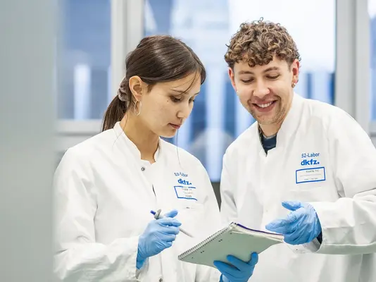 Zwei Wissenschaftler in Laboruniformen konsultieren ein Notizbuch. Die Person links, eine Frau mit langen Haaren, schaut interessiert, während der Mann rechts, mit lockigem Haar, lächelt und einen Stift in der Hand hält. Im Hintergrund sind Laborausrüstung und Fenster sichtbar, die Tageslicht hereinlassen.