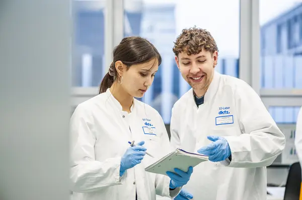 Two researchers in lab coats are engaged in discussion in a laboratory setting. One is holding a notebook and pen, while the other appears to be explaining something, both demonstrating a collaborative working environment focused on scientific research.