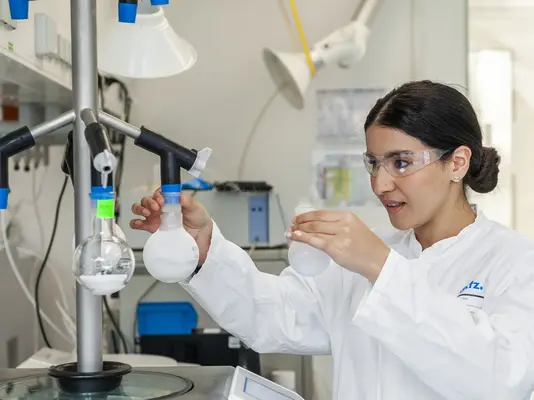 Eine Wissenschaftlerin in einem Labor trägt eine Schutzbrille und untersucht sorgfältig eine Glasflasche. Vor ihr befinden sich Laborausrüstungen und Apparaturen, die die Durchführung von Experimenten zeigen. Der Hintergrund ist hell und modern gestaltet, was auf eine professionelle Forschungseinrichtung hinweist.