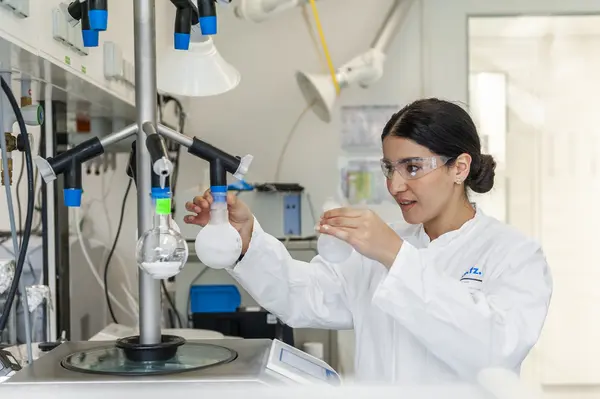 A female scientist in a lab coat and safety goggles holds a laboratory flask while setting up equipment. The laboratory is well-equipped, featuring various glassware and scientific instruments, indicating research activities taking place. The environment suggests a focus on scientific experimentation and innovation.