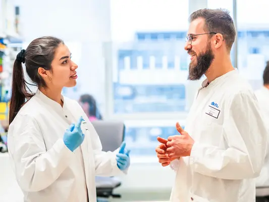 Zwei Wissenschaftler in weißen Laborkitteln unterhalten sich in einem modernen Labor. Die Frau spricht leidenschaftlich, während der Mann interessiert zuhört und gestikuliert. Im Hintergrund sind weitere Kollegen am Arbeiten zu sehen.