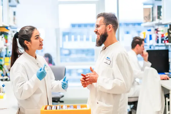 Two scientists in white lab coats engage in conversation in a bright laboratory. The woman, wearing blue gloves, gestures while talking to the bearded man. In the background, another person works at a computer, suggesting a collaborative research environment.