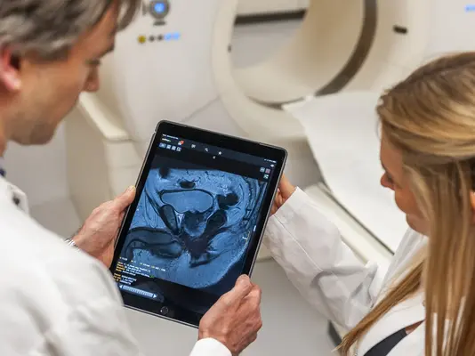Zwei medizinische Fachkräfte in weißen Kitteln betrachten gemeinsam ein Bild auf einem Tablet. Das Bild zeigt eine medizinische Untersuchung, während im Hintergrund ein CT-Scanner sichtbar ist. Die Szene vermittelt den Austausch über moderne Bildgebungstechnologien in der Diagnostik.