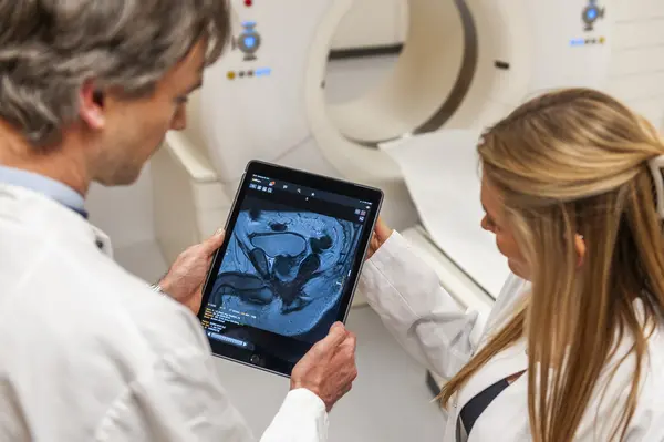 Two medical professionals are examining an MRI scan displayed on a tablet. They are in a clinical setting, with a CT scanner visible in the background. The focus is on the scan and their collaborative analysis, highlighting teamwork in medical diagnostics.
