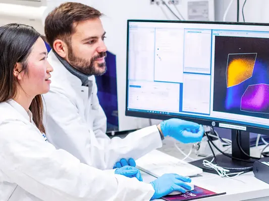 Zwei Wissenschaftler in Laborkitteln sitzen an einem Arbeitsplatz mit einem Computer. Der Bildschirm zeigt farbige grafische Datenanalysen. Beide Personen scheinen konzentriert auf die Arbeit am Computer zu sein, während sie sich gegenseitig unterstützen.