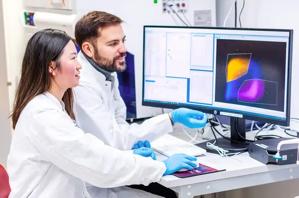 A woman and a man in lab coats are seated at a desk, focused on a computer screen displaying colorful data visualizations. The woman points at the screen, while the man observes attentively. Both wear blue gloves, and various lab equipment is visible in the background.