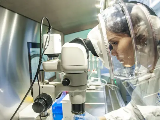 Eine Laborantin in Schutzkleidung schaut konzentriert durch ein Mikroskop. Sie arbeitet an einem Tisch mit verschiedenen Laborgeräten und Proben, um wissenschaftliche Forschung durchzuführen. Der Hintergrund zeigt eine moderne Laboreinrichtung.