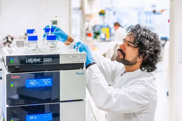A researcher in a lab coat is carefully setting up a scientific instrument, the UltiMate 3000. The equipment has several bottles with blue caps and connected tubes, indicating preparation for an experiment in a laboratory environment.