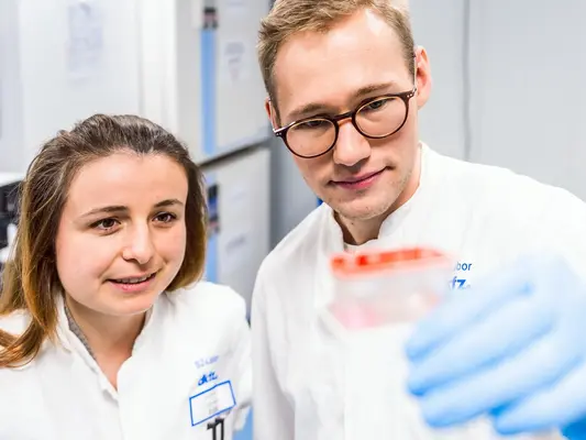 Zwei Labormitarbeiter, eine Frau und ein Mann, betrachten gemeinsam eine transparente Probenbehälter in einem modernen Labor. Die Frau trägt einen weißen Laborkittel und hat lange, halblange Haare. Der Mann trägt eine Brille und hat kurze Haare. Beide scheinen konzentriert und interessiert.