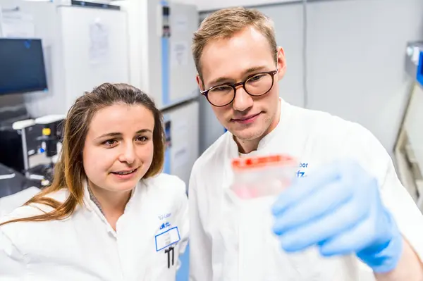 Two laboratory researchers, a man and a woman, are closely examining a petri dish. The woman, wearing a lab coat, looks intently at the dish, while the man holds it up, showing its contents. They are in a modern lab environment, highlighting collaborative scientific work.