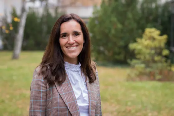 Eine lächelnde Frau mit langen, braunen Haaren steht im Freien vor einer grünen Hintergrundlandschaft. Sie trägt ein kariertes Blazerjackett über einer weißen Bluse.