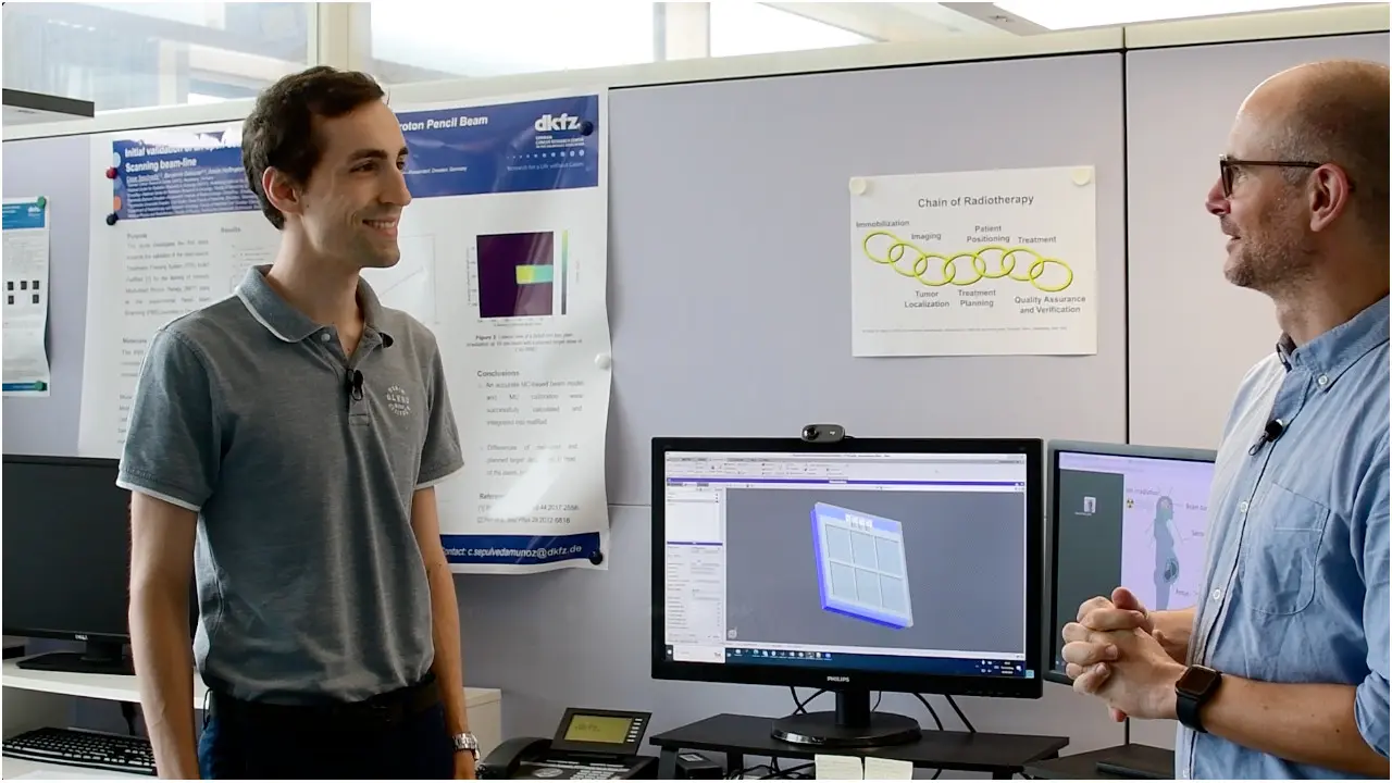 Two men are engaged in conversation in an office setting. One man, wearing a gray shirt, is smiling and listening, while the other, wearing glasses, is speaking. A computer with a design on the screen is visible between them, along with posters on the wall discussing various topics.