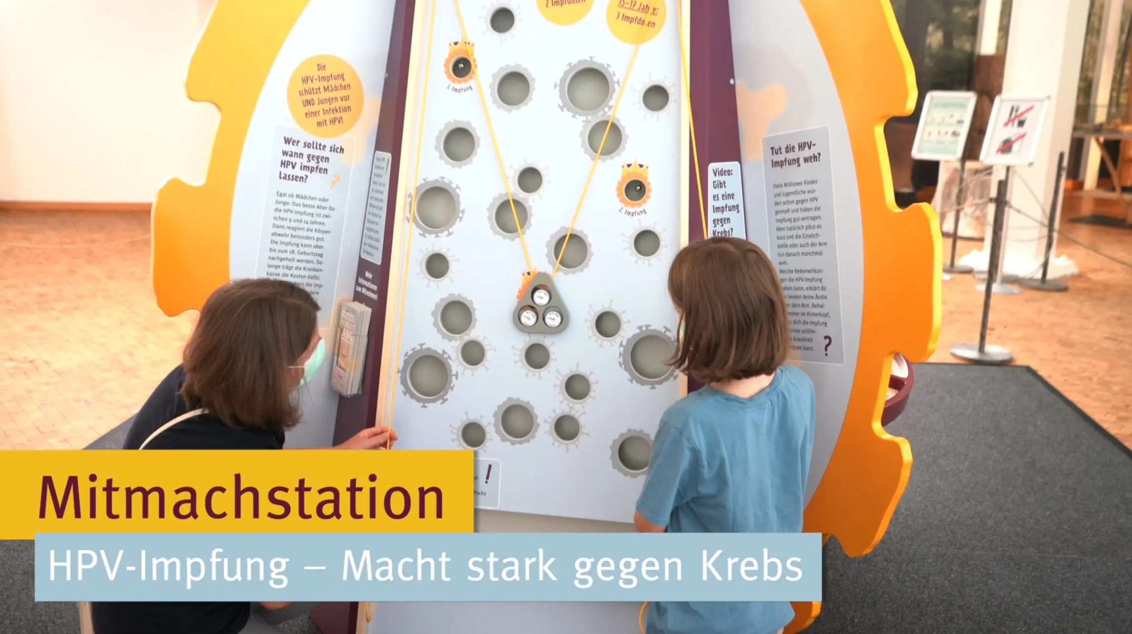 Eine Mitmachstation zur HPV-Impfung mit der Überschrift „Macht stark gegen Krebs“. Zwei Personen betrachten interaktive Elemente. Informative Texte unterstützen das Lernen über den Impfstoff und seine Bedeutung für die Krebsprävention.