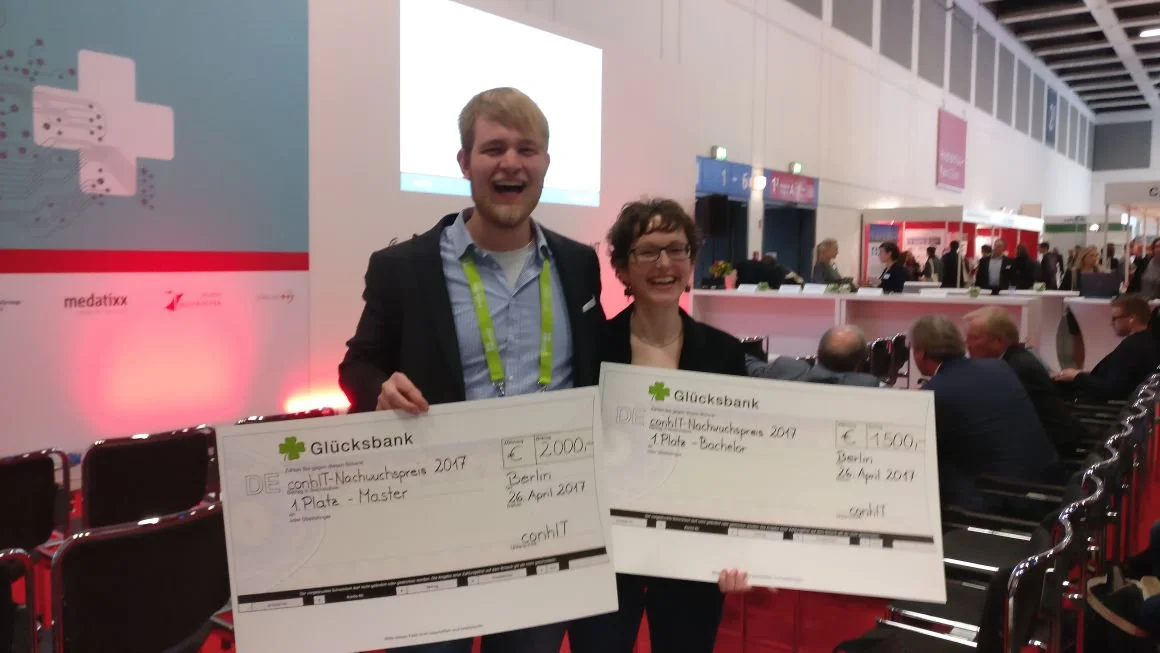 Two smiling individuals hold large ceremonial checks, celebrating their achievements at an event. The checks indicate prize amounts and recognition for their work. The background features a conference setting with various attendees and banners, suggesting a lively atmosphere.