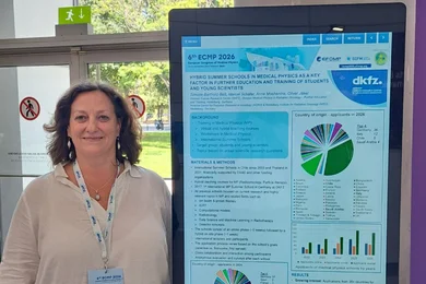 Eine Frau steht neben einem Informationsplakat auf einer Konferenz. Das Plakat enthält graphische Darstellungen und Texte zu einem wissenschaftlichen Thema, erkennbar durch Diagramme und Statistiken. Im Hintergrund sind Fenster und Bäume sichtbar.