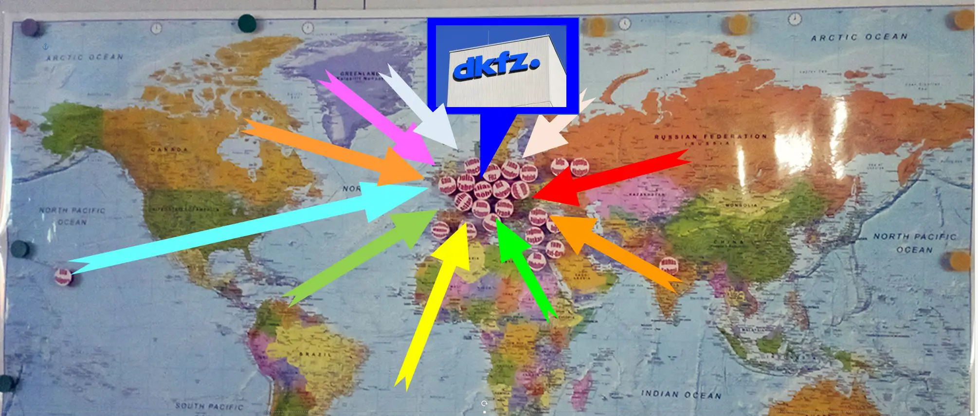 A world map featuring various locations marked with round labels, each connected by colored arrows pointing toward a central logo labeled "dkfz." The arrows indicate focus points related to the labeled sites on the map.