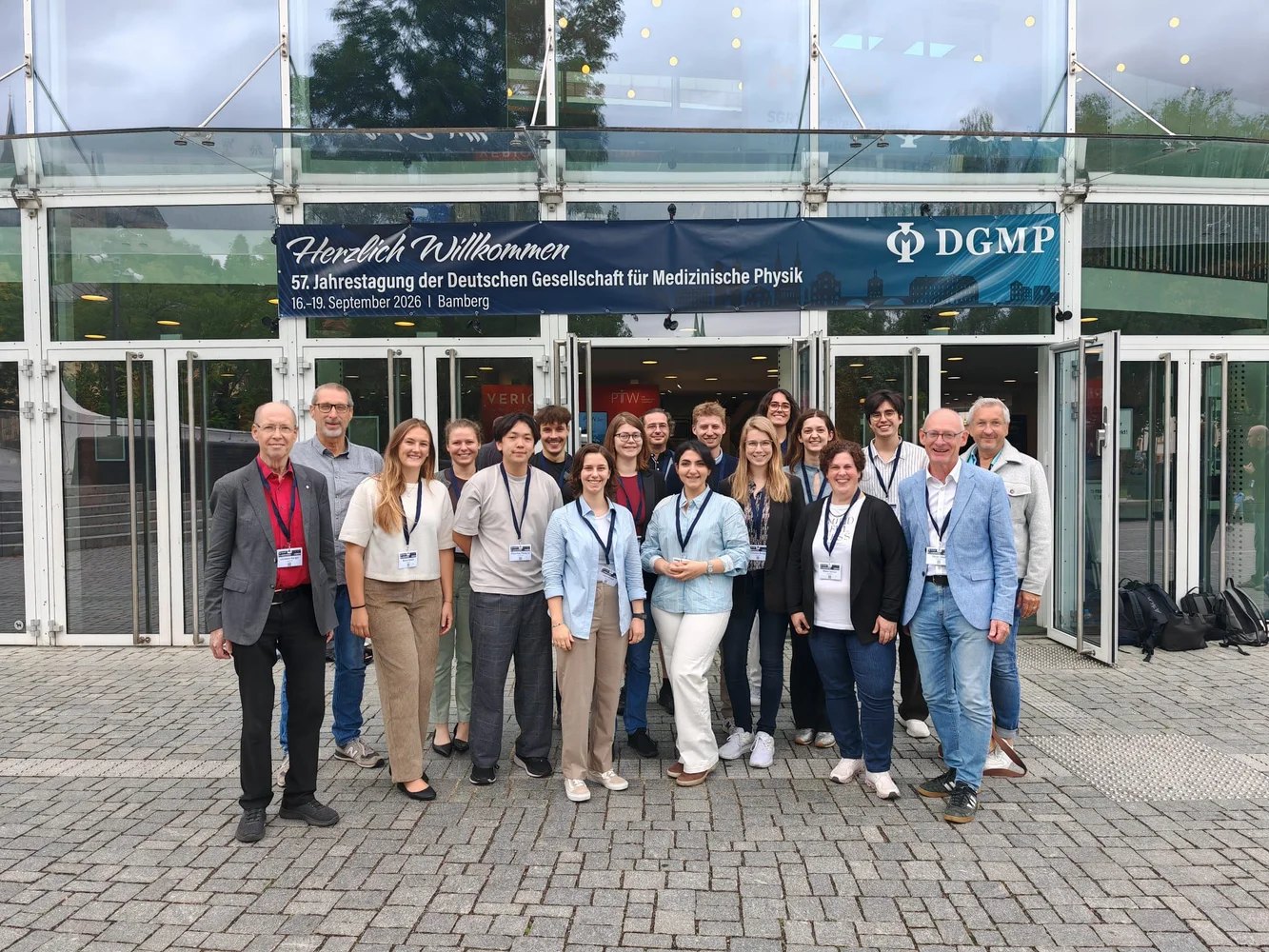 Gruppe von 18 Personen vor einem Gebäude, das eine Veranstaltung zur medizinischen Physik ankündigt. Die Teilnehmer sind unterschiedlichen Alters und Geschlechts, tragen Namensschilder und stehen auf einem gepflasterten Platz. Im Hintergrund ist ein Banner mit dem Schriftzug "Herzlich Willkommen" zu sehen.