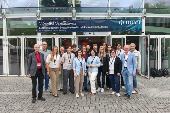 Gruppe von 18 Personen vor einem Gebäude, das eine Veranstaltung zur medizinischen Physik ankündigt. Die Teilnehmer sind unterschiedlichen Alters und Geschlechts, tragen Namensschilder und stehen auf einem gepflasterten Platz. Im Hintergrund ist ein Banner mit dem Schriftzug "Herzlich Willkommen" zu sehen.