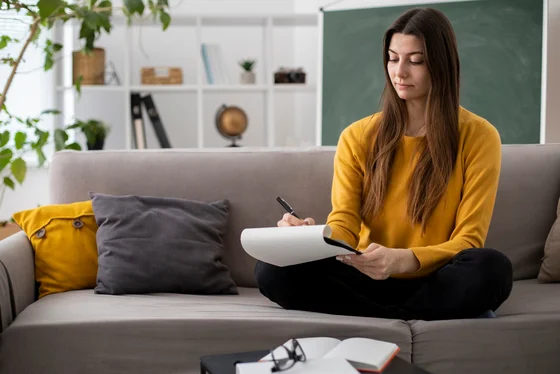 Eine junge Frau sitzt mit gekreuzten Beinen auf einem Sofa und schreibt in ein Notizbuch. Sie trägt ein gelbes Oberteil und hat lange, braune Haare. Im Hintergrund sind Regale mit Pflanzen und Büchern sowie eine Tafel sichtbar.