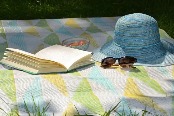 Ein blauer Sonnenhut liegt neben einer Sonnenbrille und einem aufgeschlagenen Buch auf einer bunten Decke im Grünen. Im Hintergrund ist rasengrün zu sehen. Diese Szene vermittelt eine entspannte Sommeratmosphäre im Garten, ideal für Erholung und Sonnenschutz.