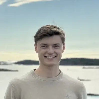 Ein junger Mann steht lächelnd auf einer Felslandschaft am Wasser. Er trägt einen hellen Pullover und schwarze Hosen. Der Hintergrund zeigt den Himmel und das Wasser, umgeben von weiteren Felsen und etwas Vegetation.