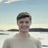 Ein junger Mann steht lächelnd auf einer Felslandschaft am Wasser. Er trägt einen hellen Pullover und schwarze Hosen. Der Hintergrund zeigt den Himmel und das Wasser, umgeben von weiteren Felsen und etwas Vegetation.
