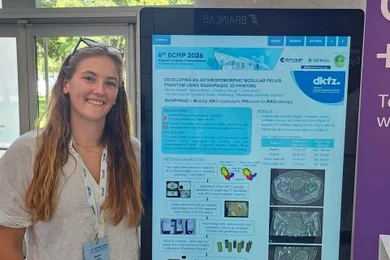 Eine Person steht neben einem Informationsdisplay, das ein wissenschaftliches Plakat zeigt. Das Plakat enthält Diagramme und Texte zu einem Thema in der biomedicalen Forschung. Hintergrund mit Pflanzen und einem Eingangsbereich zu sehen.