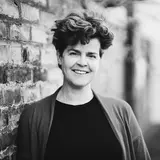 Eine lächelnde Frau mit lockigem, braunem Haar steht vor einer Steinmauer. Sie trägt einen grauen Cardigan über einem schwarzen Oberteil.