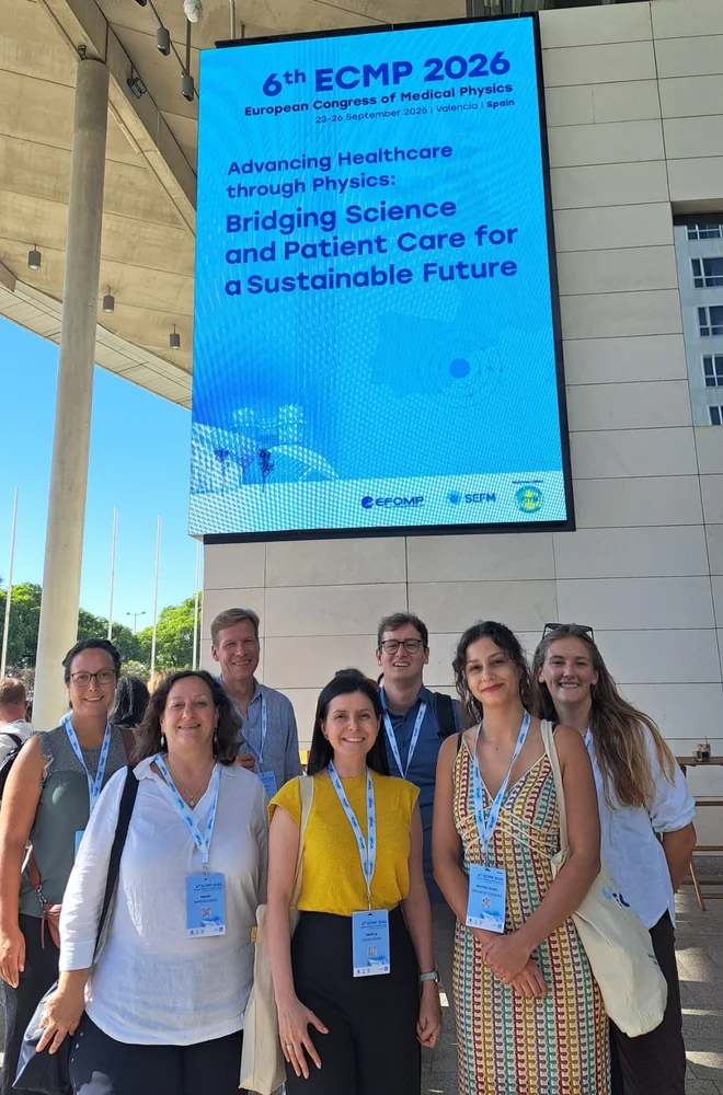 Eine Gruppe von sieben Personen steht lächelnd vor einem großen digitalen Banner, das Informationen über den 6. Europäischen Kongress für Medizinische Physik 2026 zeigt. Das Banner betont die Themen Gesundheitsversorgung durch Physik und die Verbindung von Wissenschaft und Patientenversorgung.