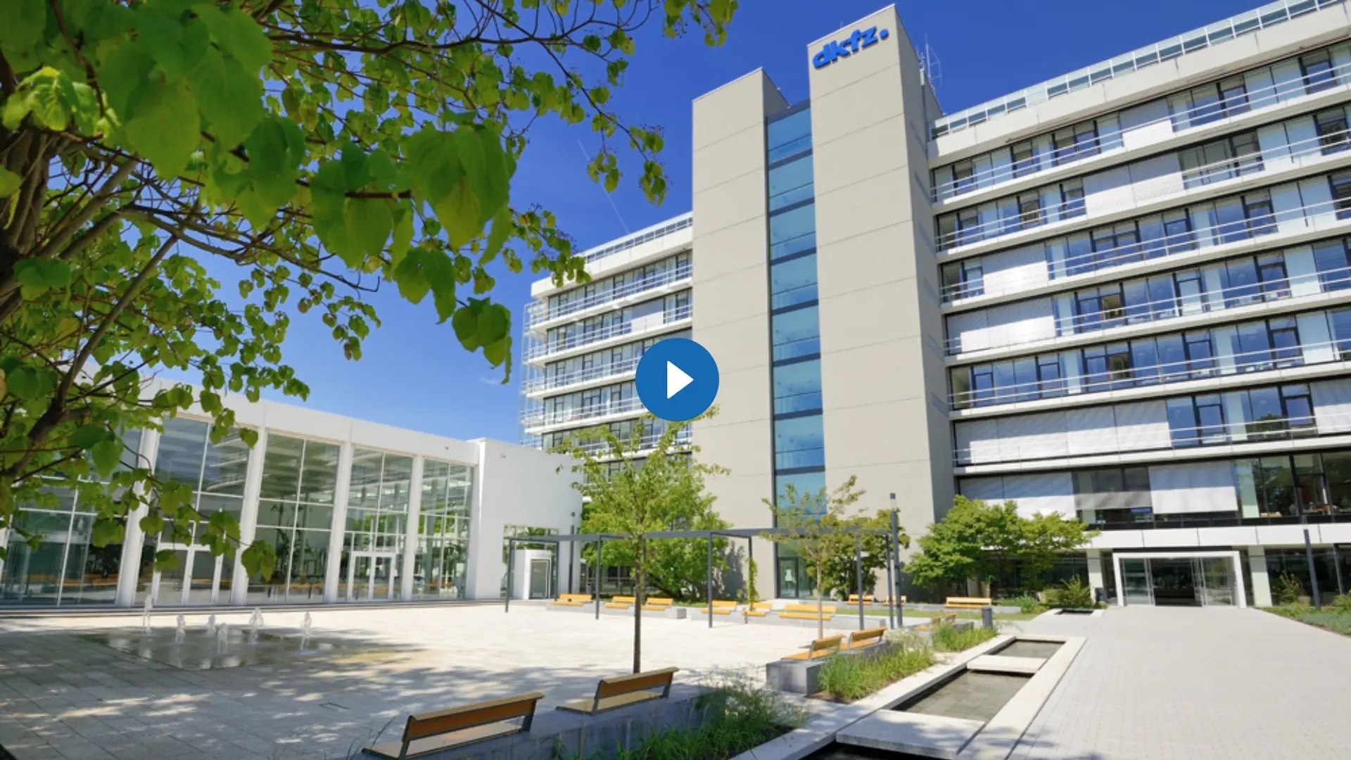 A modern, multi-story building with large glass windows is set against a clear blue sky. In the foreground, there are benches and greenery, creating a welcoming outdoor space. A play button symbol indicates a video related to the building.