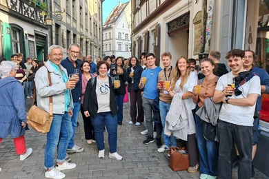 Eine Gruppe von Menschen steht in einer belebten Straße und hält Gläser mit Getränken hoch. Sie lächeln und posieren für ein Foto. Im Hintergrund sind historische Gebäude und eine Menschenmenge zu sehen.
