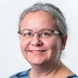 Ein Porträt einer lächelnden Frau mit kurzen, grauen Haaren. Sie trägt eine Brille und ein Oberteil mit einem blauen Muster. Der Hintergrund ist hell und neutral. Die Frau strahlt Freundlichkeit und Offenheit aus.