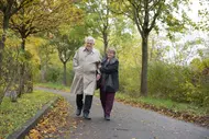 Ein älteres Paar spaziert auf einem Weg in einem Park, umgeben von herbstlichen Bäumen und buntem Laub. Der Mann trägt einen beigen Trenchcoat und die Frau einen dunklen Mantel. Sie halten sich am Arm und lächeln einander an.