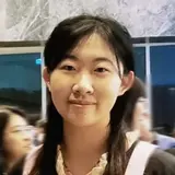 Eine junge Frau steht in einem formellen Outfit, das einen schwarzen Abschlussmantel mit pinken Bändern und einer großen, hellen Schleife beinhaltet. Sie lächelt in die Kamera und befindet sich in einer Menschenmenge, die im Hintergrund nicht klar zu erkennen ist.