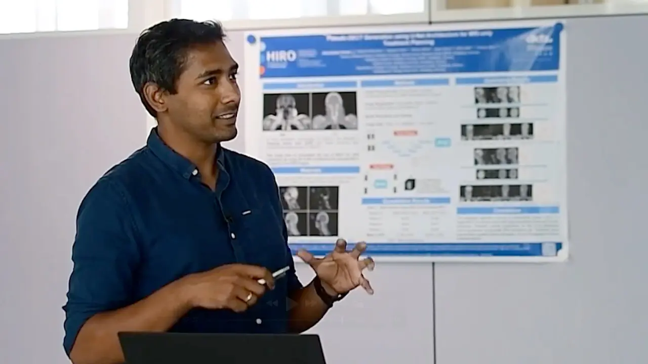 A man in a blue shirt is gesturing while giving a presentation, standing in front of a poster detailing research findings. He appears engaged and is using a pen to emphasize points. The poster includes images and text related to the research topic.