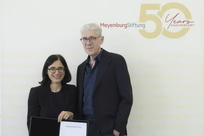 Zwei Personen stehen vor einem Hintergrund mit dem Text "Meyenburg Stiftung 50 Years Anniversary". Die Frau links hält ein Zertifikat und lächelt, während der Mann rechts neben ihr, in einem dunklen Anzug, ebenfalls lächelt.