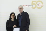 Zwei Personen stehen vor einem Hintergrund mit dem Text "Meyenburg Stiftung 50 Years Anniversary". Die Frau links hält ein Zertifikat und lächelt, während der Mann rechts neben ihr, in einem dunklen Anzug, ebenfalls lächelt.