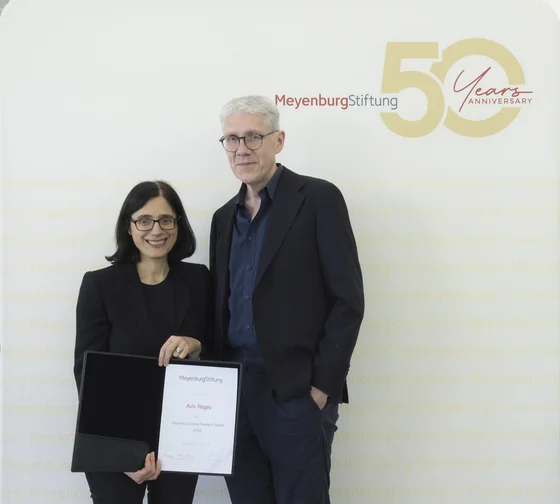 Zwei Personen stehen vor einem Hintergrund mit dem Text "Meyenburg Stiftung 50 Years Anniversary". Die Frau links hält ein Zertifikat und lächelt, während der Mann rechts neben ihr, in einem dunklen Anzug, ebenfalls lächelt.