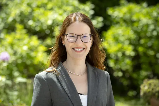 A smiling woman with long, wavy brown hair is wearing glasses and a gray blazer, standing in front of a lush green background. She appears confident and approachable. This image is associated with recent news regarding her work or achievements, emphasizing her professional presence.