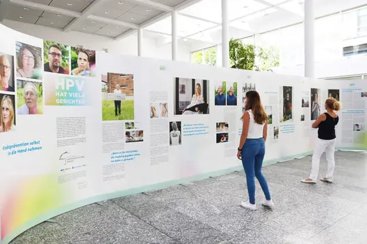 Zwei Frauen betrachten die Ausstellung ‚HPV hat viele Gesichter‘ mit Fotos und Erfahrungsberichten auf weißen Stellwänden in heller Halle.