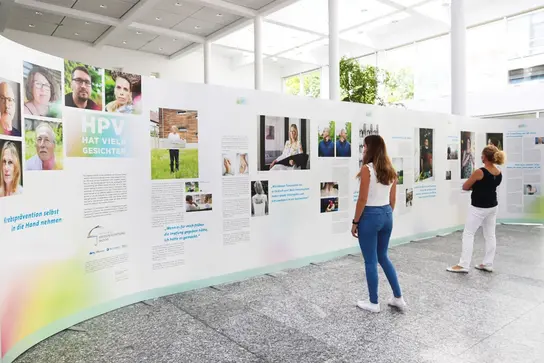 Zwei Frauen betrachten die Ausstellung ‚HPV hat viele Gesichter‘ mit Fotos und Erfahrungsberichten auf weißen Stellwänden in heller Halle.