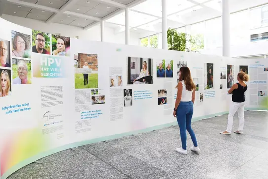 Zwei Frauen betrachten die Ausstellung ‚HPV hat viele Gesichter‘ mit Fotos und Erfahrungsberichten auf weißen Stellwänden in heller Halle.