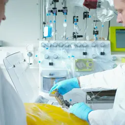 Zwei Laboranten in weißen Kitteln arbeiten in einem modernen Labor. Ein Mann bedient ein Gerät, während er eine gelbe Folie in den Händen hält. Im Hintergrund sind technische Geräte und Instrumente sichtbar, die für wissenschaftliche Experimente verwendet werden.