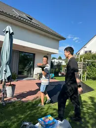 Zwei Personen stehen im Garten eines Hauses. Eine Person trägt eine große Pizza, während die andere daneben geht. Im Hintergrund sind ein Trampolin und eine grüne Hecke sichtbar. Der Himmel ist blau mit wenigen Wolken.