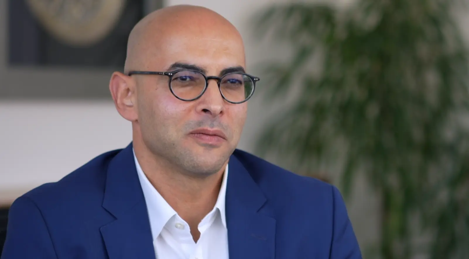 Ein Mann mit Glatze und Brille sitzt in einem hellen Raum. Er trägt einen blauen Anzug und ein weißes Hemd. Der Hintergrund zeigt eine Pflanze und eine unbestimmte Wanddekoration. Der Ausdruck des Mannes ist ernst und nachdenklich.