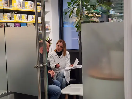Zwei Personen sitzen in einem modernen Beratungsraum. Eine Frau in einem weißen Kittel bespricht Dokumente mit einem Mann. Im Hintergrund sind Bücherregale mit Informationsmaterialien zu sehen.